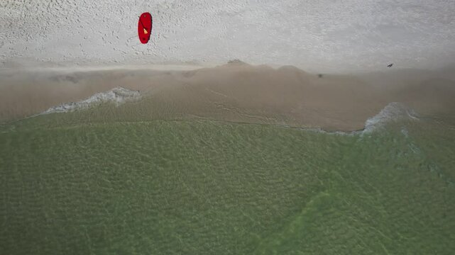 Aerial view of a beach with a surfboard, and red objects on the sand, next to the shoreline with gentle waves.