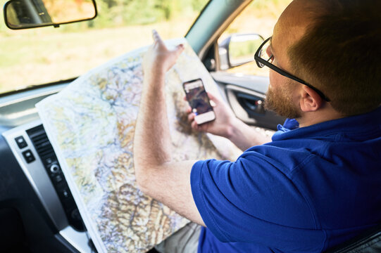 Man tourist sits in passenger seat of car, holding map and smartphone with navigation app. Traveler points out of windshield with focused expression, planning his route or identifying landmark ahead.