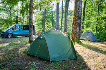 Multiple green tents in wooded campsite, with blue van parked nearby. Orange hammock strung between two trees. Sun filters through trees, casting dappled light across serene and inviting campsite.