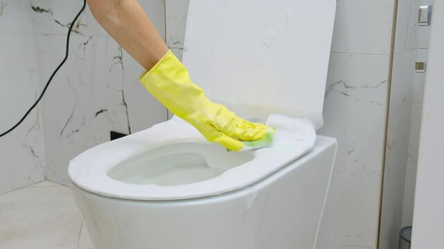 cleaning toilet seat restroom at house green sponge rag Keeping house neat , female wearing yellow rubber gloves Housekeeper spray liquid cleaner foam toilet lid disinfecting with disinfectant 