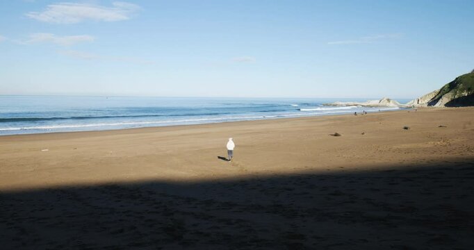 Mujer andando por la playa, pasando de la zona en sombra a la zona soleada, en los primeros d&iacute;as de primavera, plano general.