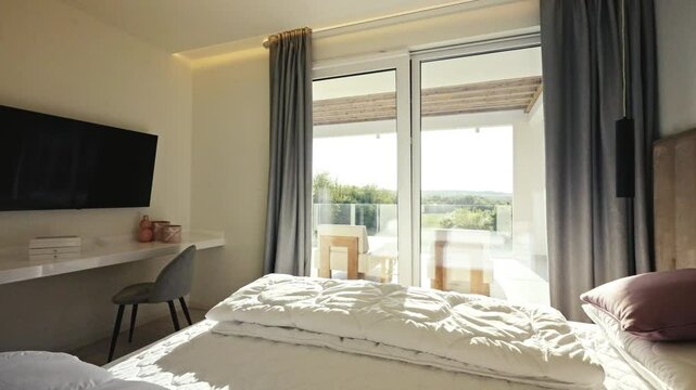 Bright and elegant bedroom interior with a cozy bed, modern TV setup, and a balcony overlooking a lush, green landscape. Concept of luxury accommodation and relaxing spaces