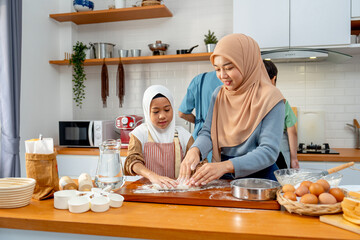 Young Asian muslim mother help and teach her daughter to threshing flour by hands on wooden tray for making the bakery with father and son also cook in the back in the kitchen of their house.