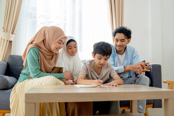 Muslim family with mother, father and daughter sit and cheer little boy for writing in living room of their house.