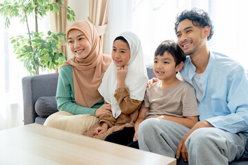 Asian muslim family compose of young mother, father, daughter and son hug and stay together on sofa in living room of their house and look forward and right direction of image with smiling.