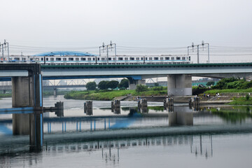 A Shinkansen train crosses a bridge over a calm river, reflecting its structure in the water, surrounded by green banks and distant industrial areas of Tokyo.