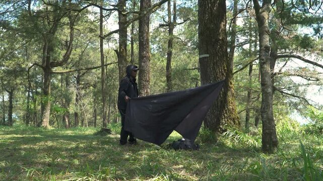 A solo traveler is preparing a flysheet tent in a green and lush pine forest area. Asian man adventuring alone in the jungle and setting up a trap tent from flysheet for bushcraft and camping