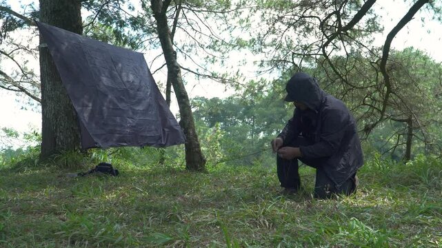 A solo traveler is preparing a flysheet tent in a green and lush pine forest area. Asian man adventuring alone in the jungle and setting up a trap tent from flysheet for bushcraft and camping