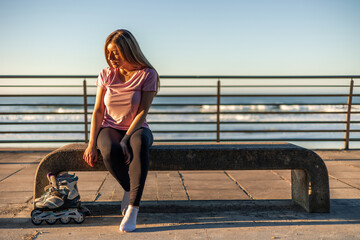 Young woman resting on bench after inline skating by the sea at sunset copyspace