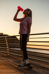 vertical Sporty woman drinking water and wearing inline skates at sunset