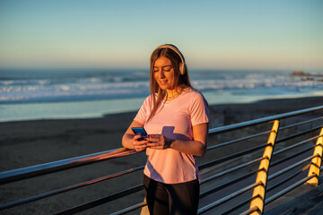 Sportswoman using smartphone and listening music with headphones at sunset on the beach