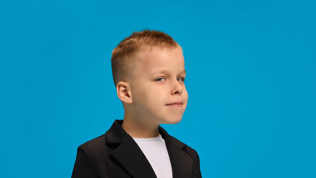 Boy wearing black blazer and white shirt, glancing sideways with mischievous smile, exuding playful and sly energy against bright blue background. Concept of childhood, beauty, fashion, style. Ad
