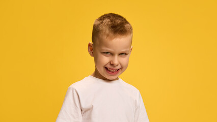 Portrait of boy showing wide mischievous grin, tilting head forward slightly, wearing white t-shirt against yellow background. Concept of childhood, beauty, fashion, style. Ad