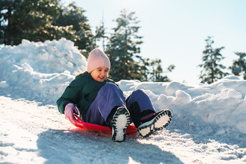 Happy girl smiling while sledding down a snowy slope on a vibrant red plastic sled. Enjoying a...