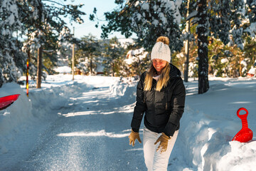 Smiling young woman in a black puffer jacket and white pants throws snow in the air while playing...