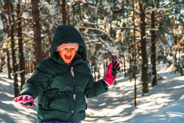 Happy child wearing a warm hooded winter jacket and pink gloves is playing with snow in a beautiful winter forest during a sunny winter day