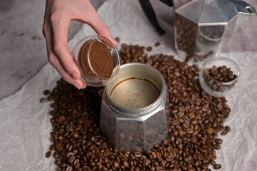 Woman's hands pouring coffee from a small transparent cup in geyser coffee maker moka machine. Scattered fresh aromatic coffee beans around. Light background. Preparation to making coffee