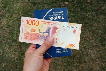 Person holding brazilian passport and 1000 pesos argentinos. Brazilians travelling to Argentina concept. 