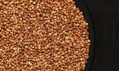 Raw buckwheat in a black plate on a dark wooden table. Buckwheat groats in a plate, top view. Raw buckwheat grains, uncooked porridge. Buckwheat background
