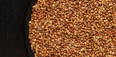 Raw buckwheat in a black plate on a dark wooden table. Buckwheat groats in a plate, top view. Raw buckwheat grains, uncooked porridge. Buckwheat background