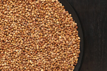 Raw buckwheat in a black plate on a dark wooden table. Buckwheat groats in a plate, top view. Raw buckwheat grains, uncooked porridge. Buckwheat background