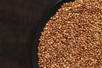 Raw buckwheat in a black plate on a dark wooden table. Buckwheat groats in a plate, top view. Raw...