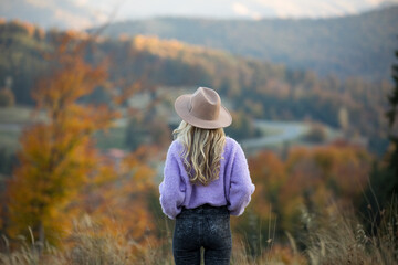 Blonde woman wearing a hat and sweater stands in a vibrant autumn landscape with colorful trees and distant mountains, evoking a sense of exploration, solitude, and the beauty of the fall season.