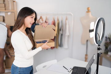  A woman enthusiastically livestreams while holding a box, showcasing her products with a ring light and laptop setup in a workspace filled with racks and packaging
