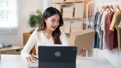 Fototapeta premium A woman enthusiastically livestreams while holding a box, showcasing her products with a ring light and laptop setup in a workspace filled with racks and packaging