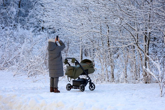 Young woman with a baby stroller walking in winter park. Snow weather, mom with pram photograph the nature on smartphone camera
