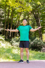 Sporty happy Japanese guy practicing yoga meditation, receives positive energy, healthy lifestyle. Young man standing making breath exercises, relaxing in park on sunny summer day. Active sportsman.