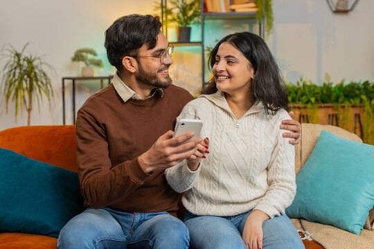 Multiracial young couple sitting on sofa and swipe smartphone at home. Happy Arabian man and woman discussing together spend time, relaxing and enjoying communicate on mobile phone in room apartment