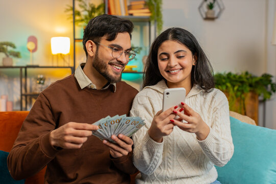 Happy exited Indian couple holding money cash and smartphone planning discussing investment together sitting on sofa in living room at home. Successful smiling family after winning lottery jackpot
