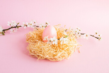 Pink easter egg in a nest of straw and white flowering tree branches on the pink background....