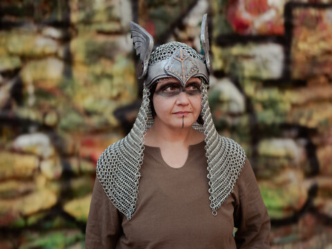 Female Viking in winged and chainmail armor and face paint against graffitied wall. 
