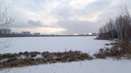 Winter landscape with frozen river, trees and residential buildings in the distance