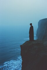 Fototapeta premium Silhouetted figure standing on a cliff overlooking a foggy ocean.