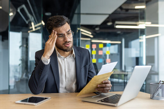 A Latin American businessman sitting in a sleek office, studying documents with a concerned expression, suggesting financial or business-related challenges, using a laptop for analysis