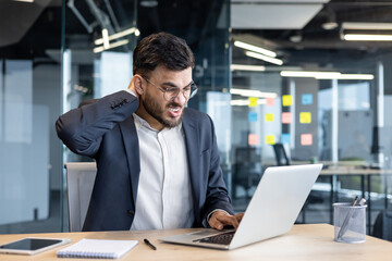 Fototapeta premium Latin American businessman in a modern office experiencing neck discomfort working diligently on a laptop, reflecting a moment of workplace stress or computer-related strain during a busy workday.