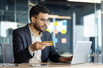 Latin American businessman sitting at desk in office, conducting online transactions on laptop with a credit card in hand, highlighting modern business and technology.