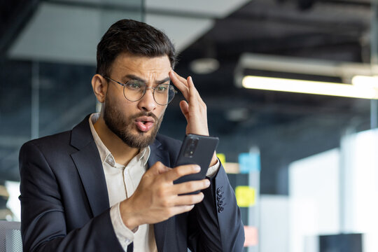 A Latin American businessman in a suit expresses shock while looking at his smartphone in an office.