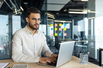Latin American businessman focused on his work while using a laptop in a bright, professional office environment, embodying dedication, focus, and professionalism in a corporate setting.