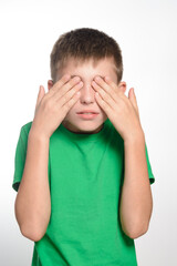 A young boy with short brown hair is shown covering his eyes with his hands. He's wearing a bright green t-shirt against a plain white background. The image conveys a sense of playfulness or shyness.