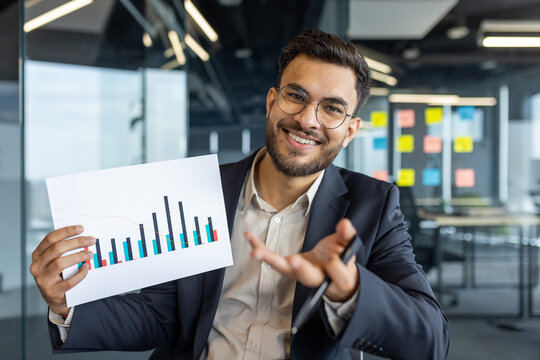 Latin American businessman holding a report with financial charts, presenting data in a modern office with enthusiasm and professionalism, conveying success, business growth, and strategic planning
