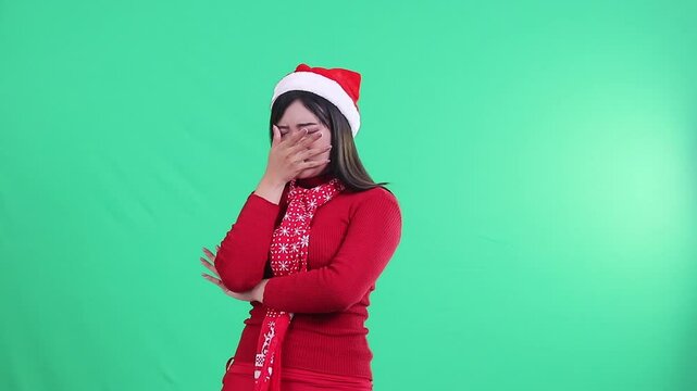 Asian girl wearing Christmas sweater, Santa hat and scarf, showing expression of smelling bad smell while looking nauseous on green background, green screen. Suitable for creative projects