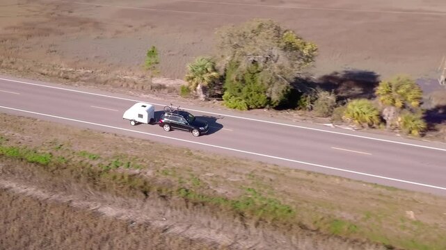 Aerial tracking view of vehicle pulling teardrop camper along coastal highway.