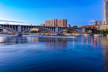 Naklejka premium City skyline reflected on tranquil river at dusk, under a bridge. Urban landscape. Mississippi River, Minneapolis, Minnesota, United States