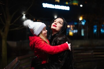 Smiling mother and daughter enjoy the winter evening in a festive urban park surrounded by lights
