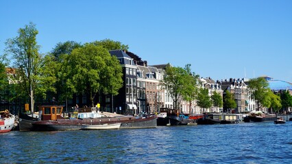 Charming Amsterdam canal with houseboats moored along the waterfront, lined by historic black and brick townhouses, lush green trees, and a vibrant blue sky completing the serene scene