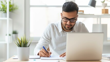 Obraz premium A focused man works at a laptop while taking notes, surrounded by a plant and bright natural light in a modern workspace.